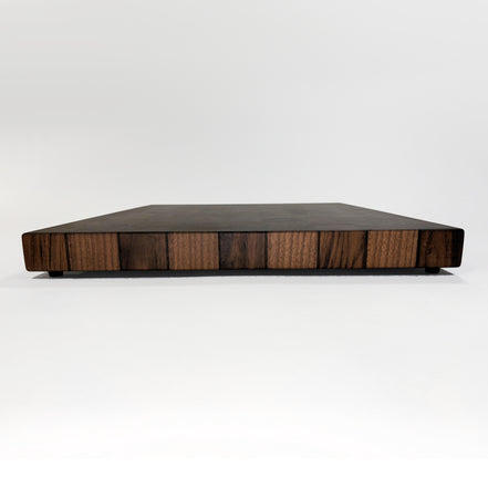 A rectangular wooden cutting board with a dark top surface and a striped wood-grain patterned side, photographed on a seamless white studio background.