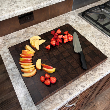 A dark brown walnut wood basket weave cutting board on a granite kitchen countertop, shown with sliced apples, strawberries, and a chef's knife.